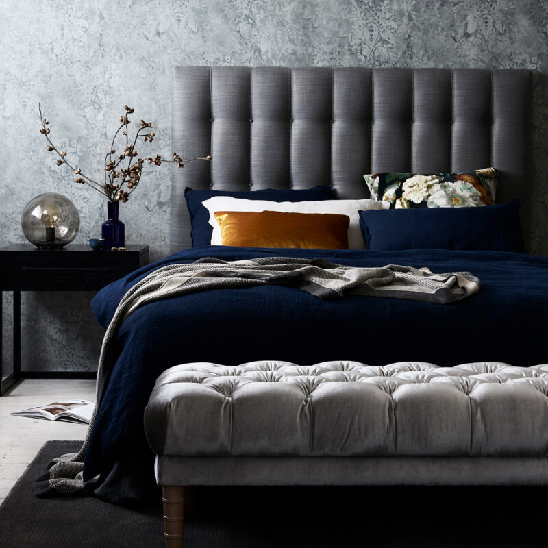 A tufted gray headboard against a textured blue wall, with layered dark blue and patterned pillows on a neatly made bed.