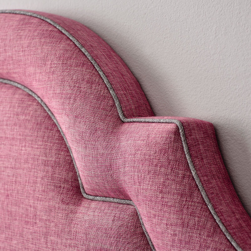 Close-up of a pink upholstered headboard with an arched design and contrasting gray piping against a neutral wall, conveying elegance and comfort.