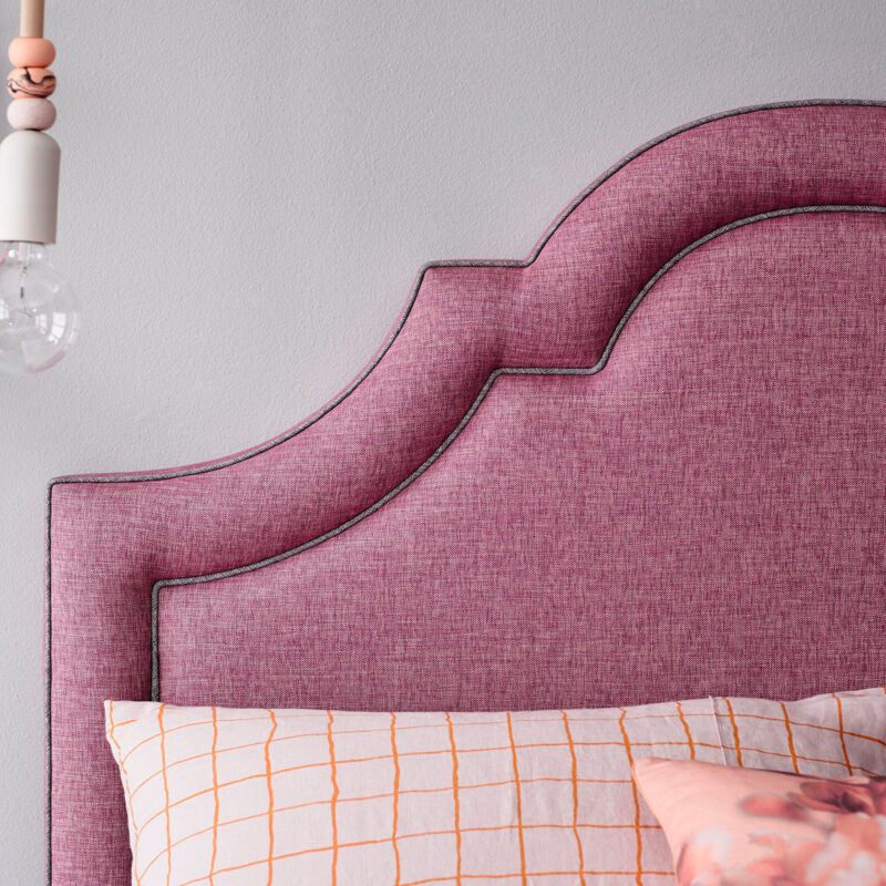 Plush pink headboard with elegant curves against a gray wall, beside a light bulb fixture. A pillow with an orange grid pattern adds contrast.