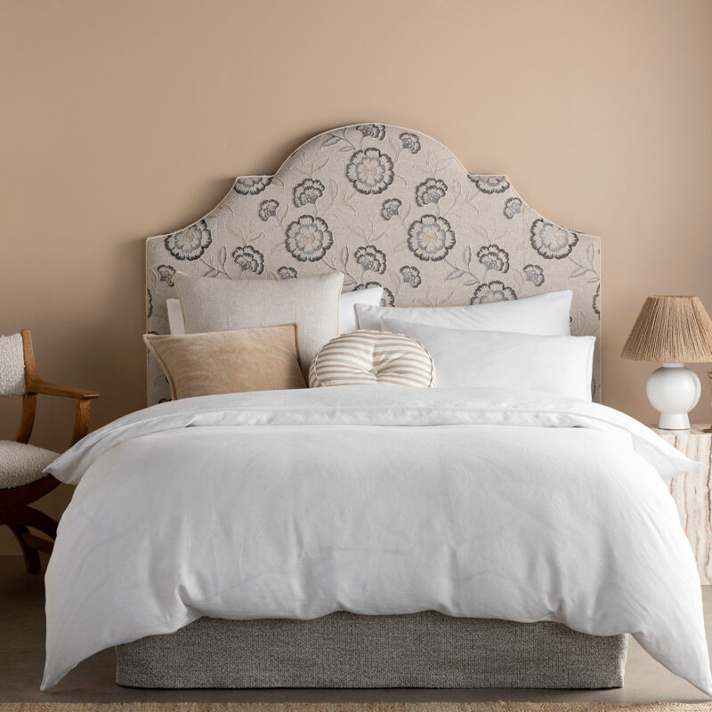 Cozy bedroom featuring a floral upholstered headboard, layered white bedding, and soft pillows, with a stylish chair and bedside lamp.